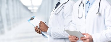 Tıbbi destek. Hastanenin koridorunda elinde pano ve dijital tabletle dikilen iki erkek doktorun görüntüsü. Üniformalı tanınmayan terapistler hasta ve kolaj muayenesine hazır.