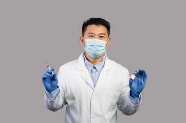 Beyaz önlüklü, koruyucu maskeli, ciddi bir Çinli terapist gri arka planda aşı ve şırınga gösteriyor. Sağlık hizmetleri için aşı, covid-19 için tıbbi tedavi