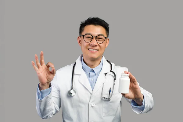 身穿白衣的快乐的中国成年男性治疗师 眼镜上有一罐药丸和手签 与灰色背景隔离 医生对治疗 保健药品的建议 — 图库照片