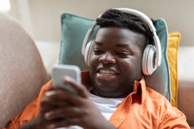 Bin yıllık tombul siyahi bir adamın cep telefonu ve kablosuz kulaklıkla gündelik koltukta uzanmış gülümseyerek internetten içeriği izlerken güzel bir uygulamanın keyfini çıkarırken çekilmiş yakın plan fotoğrafı.