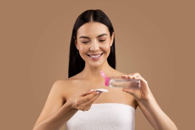 Gülümseyen, yarı çıplak, banyo havlusuna sarılı, bej rengi stüdyo arka planında izole edilmiş su ve pamuk pedi kullanan, yüzünü temizleyen, makyajını silen, uzay fotokopisi çeken güzel bir kadın.