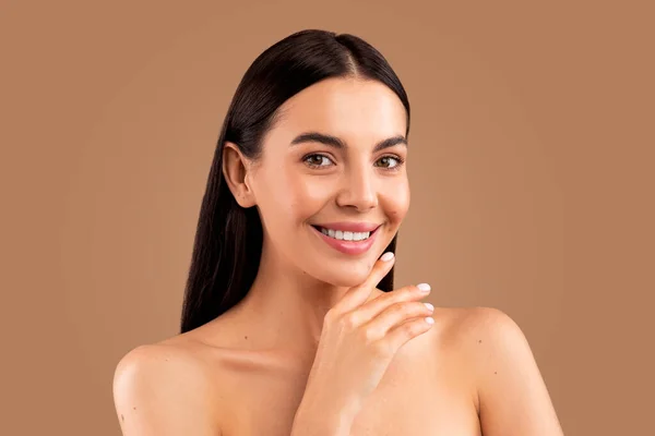 Aesthetic medicine and cosmetic surgery concept. Headshot of attractive long-haired brunette half-naked lady demonstrating her young smooth skin, touching face and smiling at camera, beige background
