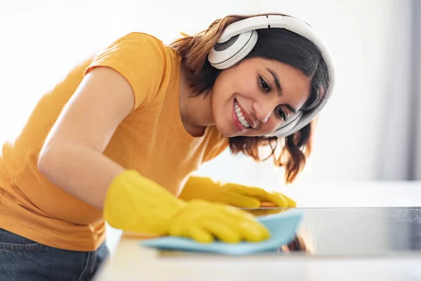 キッチンカウンターを掃除しながら音楽を聴いている陽気な若いアラブ人女性 無線電話やゴム手袋を身に着けている中東の女性を笑顔ぼろとテーブル表面をタイディング — ストック写真