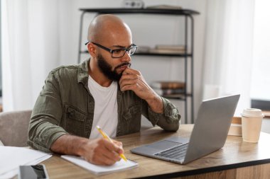 Laptop ekranına bakan, not defterine yazan, iş yerinde çalışan Latin bir adam. Konsantre erkek notlar alıyor, web sitesinde ders çalışıyor, boş alan