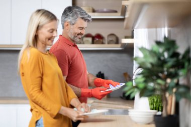 Mutlu olgun eşler birlikte ev işi yapıyor, güzel orta yaşlı beyaz saçlı adam ve sarışın kadın yemekten sonra bulaşıkları yıkıyor ve bulaşıkları yıkıyor, rahat mutfak iç mekanı, yan manzara, fotokopi odası.