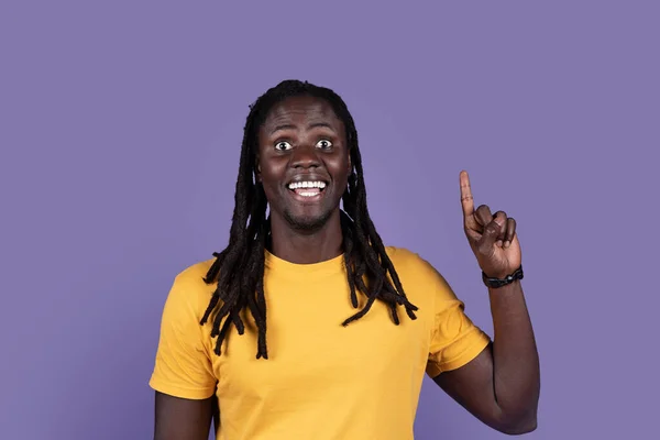黄色のTシャツを着た幸せな若い長髪のアフリカ人男性が アイデアやユーレカのジェスチャーと笑顔を示す上を向いて 興味深い計画を生み出します スタジオは紫色の背景を撮影し スペースをコピー — ストック写真