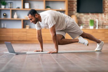 Evde dizüstü bilgisayarın önünde spor yapan atletik siyahi bir adam oturma odasının içinde yoga minderinde aktif Afrika kökenli Amerikalı erkek eğitimi alıyor, bilgisayar başında video dersleri izliyor, boş yer var.