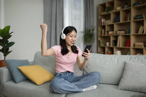 スマートフォンを見て ワイヤレスヘッドフォンで興奮した中国人女性は 彼女の手を持ち上げ 自宅で勝利のジェスチャーを作ります 若いアジアの女性は彼女の興奮を表現し ソファに座って — ストック写真