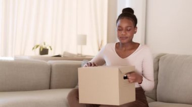 Uzun zamandır beklenen teslimat. Genç, duygusal, Afro-Amerikalı kadın karton kutuyu alışveriş siparişiyle açıyor ve evde keyif alıyor, yavaş çekim, boş alan.