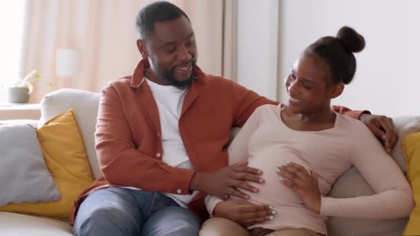 祝你怀孕愉快年轻的积极的非洲裔美国男人爱抚他怀孕妻子的大肚子 讨论她们的生儿育女问题 一起躺在沙发上 跟踪镜头 慢动作 空虚的空间 — 图库视频影像