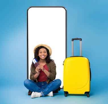 Neşeli genç siyahi kadın parlak bavulun ve büyük akıllı telefonun yanında oturuyor elinde uluslararası pasaport ve uçak biletleri, mavi stüdyo geçmişi var. Seyahat uygulaması için model ya da harika bir teklif