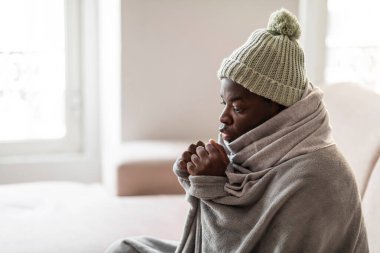 Daire ısıtma sorunu kavramı. Donmuş milenyum siyahi adamın evdeki kanepede oturup örgü ördüğü, sıcak battaniyeyle örtüldüğü, ağzının yanında el ele tutuşup ısındığı, yan gözle baktığı yakın plan.