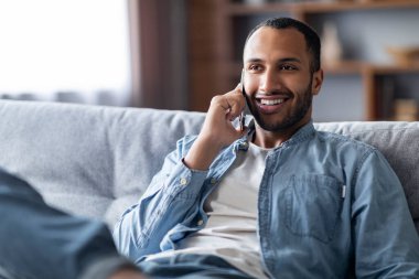 Mobil İletişim. Evde cep telefonuyla konuşan gülümseyen siyah bir adamın portresi, oturma odasında oturan yakışıklı Afrikalı Amerikalı adam ve keyifli telefon konuşmaları, yakın çekim...