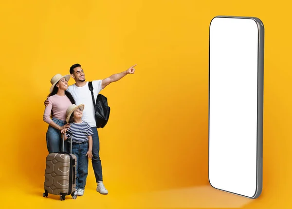 Family Travel Deal Concept. Happy middle eastern parents with little daughter carrying suitcases and pointing aside at big phone with movkup on yellow background, mom, dad and child ready for vacation