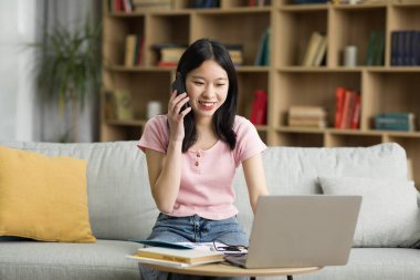 Telekomünikasyon kavramı. Bilgisayarı kullanan mutlu Asyalı kadın, akıllı telefonla konuşan, bilgisayar ve telefonla çalışan, evdeki kanepede oturan genç bayan.