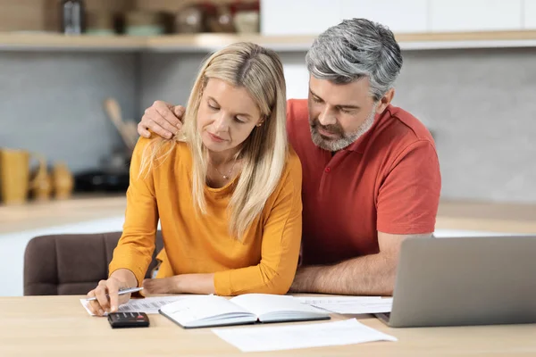 Middle aged spouses sitting at kitchen desk full of papers in front of computer, using calculator, mature man and woman counting incoming and outgoing money, planning family budget together