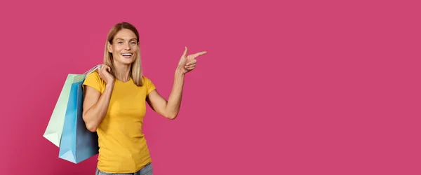令人高兴的是 成熟的高加索金发女性穿着黄色T恤 有很多包 购买手指在复制空间 孤立在粉红色的背景下 工作室 买东西 买东西 做广告 大减价 — 图库照片