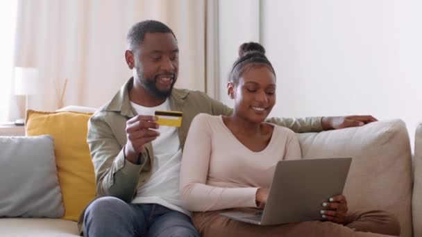 インターネット購入の快適さ 若い幸せなアフリカ系アメリカ人のカップルが一緒にオンラインショッピングを愛し クレジットカードとラップトップを使用して 自宅でソファの上に休み 撮影を追跡し 動きを遅くし 空のスペース — ストック動画
