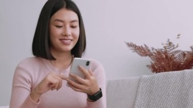 Genç, güzel Asyalı kadın internette sörf yapıyor, akıllı telefondan haberlere kayıyor ve inanılmaz reklamlar okuyor, evde dinleniyor, yavaş çekim, boş alan.