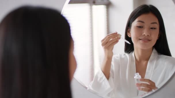 美容化妆品 年轻的亚洲女人在脸上涂营养血清 早上对着镜子的倒影微笑 慢动作 自由自在 — 图库视频影像