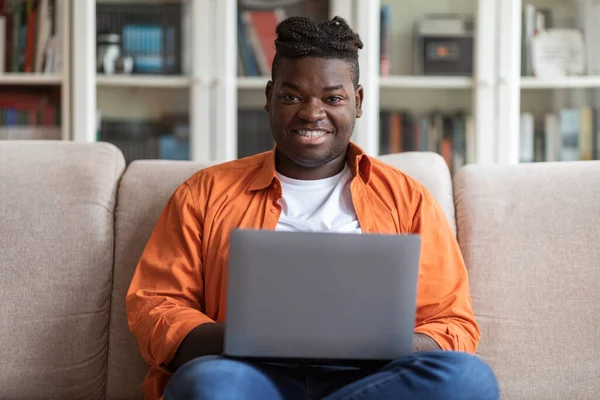 Remote job concept. Portrait of smiling happy young overweight african american guy in casual sitting on sofa, using modern laptop at home, data analyst typing on computer keyboard, copy space