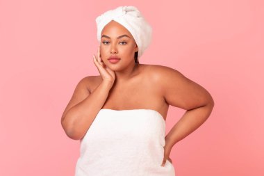 Güzel, iri yapılı, banyo havlusuna sarılı siyah bir kadının portresi, pürüzsüz ciltli güzel bir kadın kameraya bakıyor, pembe arka planda poz veriyor, stüdyo çekimi yapıyor.