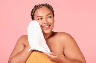 Mutlu siyah, iri yarı, temiz yüzü yumuşak beyaz havluyla kuruyan, pembe stüdyo arka planında duran kadın. Yüz temizliği ve sabah güzellik rutini