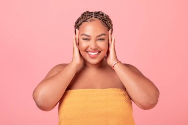 Heyecanlı siyah kadın portresi. Yüzüne dokunan, pürüzsüz teninden zevk alan, kameraya gülümseyen, pembe stüdyo arka planında poz veren pozitif kadın.