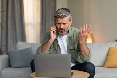 Sinirli, yaşlı, beyaz bir erkek telefonla arıyor, bağırıyor, laptopa bakıyor, el kol hareketi yapıyor, oturma odasının içindeki iş ve servisle ilgili sorunları var. Stres, çevrimiçi hata ve evdeki insan duyguları