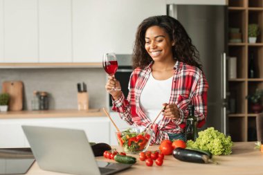 Blog ve video çağrısı. Neşeli bin yıllık siyahi kadın blogcu salata hazırlıyor, bir bardak şarap içiyor ve mutfağın içinde dizüstü bilgisayara bakıyor. Tatil kutlaması, dinlenme ve rahatlama, randevu, evde sağlık hizmeti
