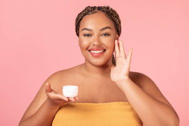Bakım konsepti. Mutlu siyah kadın yüzüne güzellik ürünü sürüyor, krem kullanıyor, havluya sarılıp pembe stüdyo arka planında poz veriyor.