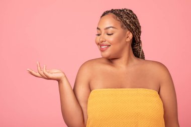 Güzel, siyah, büyük beden bir kadın elinde görünmez bir nesne, avucunun içinde duruyor, yeni bir güzellik ürünü öneriyor, pembe arka planda poz veriyor, boş alan.