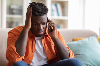 Kanepede oturan, cep telefonuyla konuşan, kafasına dokunan üzgün, şişman siyahi adamın yakın çekimi. Üzgün Afrikalı Amerikalı genç adam akıllı telefondan konuşuyor, kötü haberler var.