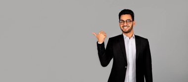 Takım elbiseli yakışıklı, gülümseyen Arap iş adamı gri stüdyo arkaplanı bir yana, genç, orta doğulu erkek girişimci iş teklifiniz için boş yer gösteriyor, panorama