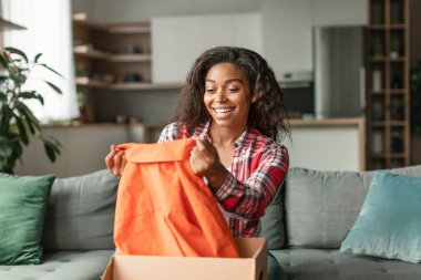 Milenyum siyahi kadın alıcının karton kutuları açması, paketin içindeki kıyafetlerin ve oturma odasındaki dağıtım hizmetlerinin keyfini çıkarması tatmin edici. Kutusuz alım, evde online alışveriş ile büyük indirim.