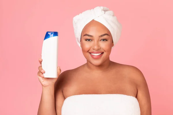 Happy black oversize lady advertising shampoo or shower gel, showing bottle and smiling to camera, pink background, mockup. Woman wearing bathrobe, recommending cosmetics