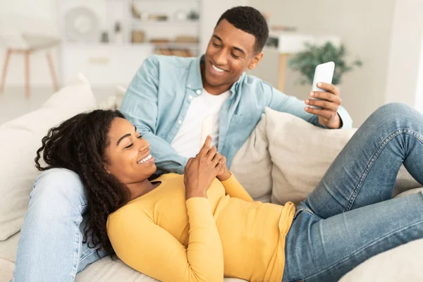 若いアフリカ系アメリカ人の男性と女性の休憩を笑顔 スマートフォンを入力し インターネットでサーフィンし リビングルームのインテリアでソファの上でビデオを見ています チャットや検索 自宅で無料でソーシャルネットワーク — ストック写真