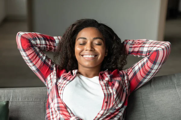 Portrait of satisfied happy millennial black lady with closed eyes relaxing on sofa, enjoying free time, comfort and peace in living room interior, close up. Weekend at home alone, rest and quiet