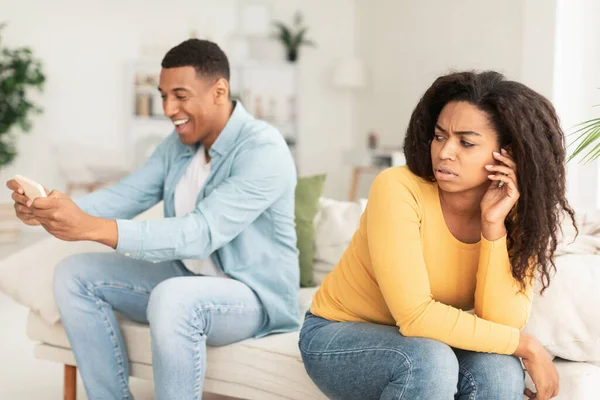 悲しい若い黒の妻は 夫を見て オンラインゲームをプレイし 部屋のインテリアでスマートフォン上のソーシャルネットワークでチャット 関係の問題 ガジェット中毒や感情 — ストック写真