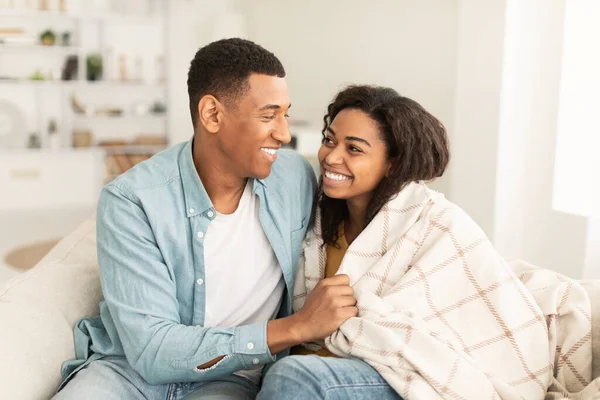 Happy young african american male covers with blanket and hugs his wife, sit on sofa, enjoy spare time in light living room interior, free space. Family love, relationship, support and care at home