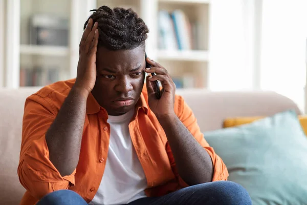 Closeup of sad overweight black guy sitting on couch, having conversation on mobile phone, touching his head. Upset african american young man talking on smartphone, having bad news
