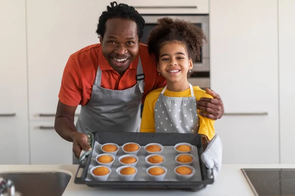 Homemade Pastry. Happy black father and his preteen daughter holding tray with baked muffins in kitchen, cheerful african american dad and female child wearing aprons and smiling at camera