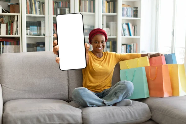 快乐的年轻黑人女人 身穿红色头巾的购物狂 头戴五颜六色的购物袋 坐在沙发上 在家里放着白色空白屏幕的手机 网上购物概念模型 自由空间 — 图库照片