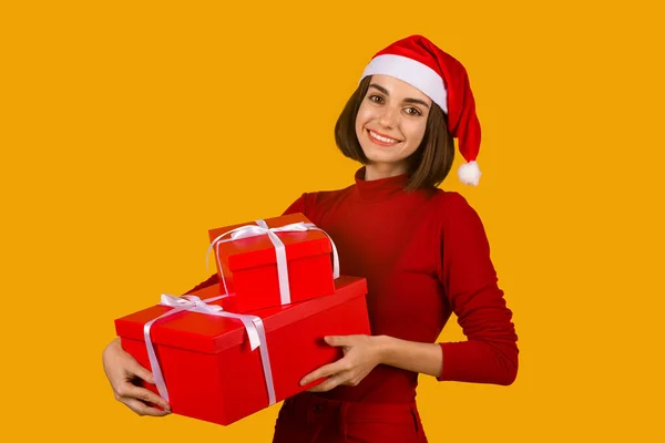 サンタハットの楽しい若いブルネットの女性と オレンジのスタジオの背景に隔離された友人や親戚のためのクリスマスプレゼントの束を保持するカジュアルな赤い衣装は スペースをコピーします クリスマスお祝いのコンセプト — ストック写真