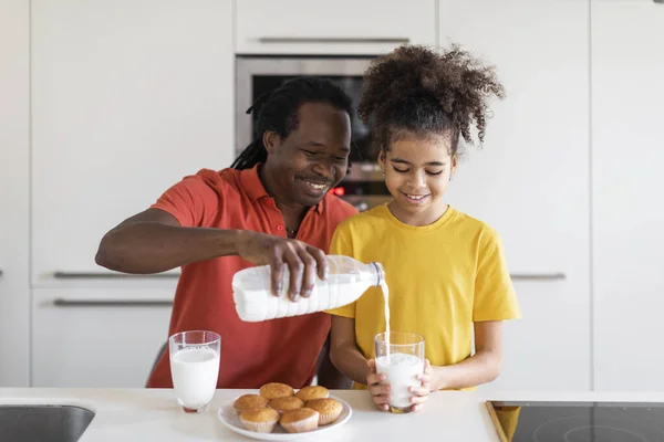Happy Black Dad And Daughter Eating Muffins And Drinking Milk In Kitchen, Smiling African American Father And Female Child Having Snacks Together, Daddy Pouring Calcium Drink From Bottle To Glass