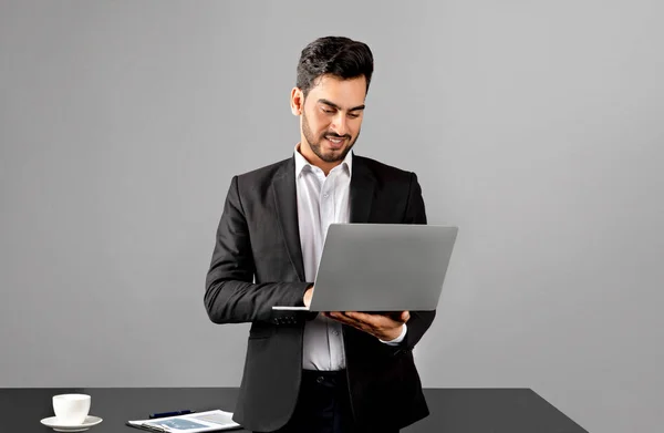 Portrait Of Smiling Arab Businessman In Suit Using Laptop In Office, Handsome Young Middle Eastern Male Entrepreneur Standing Near Desk And Using Computer For Online Communication, Copy Space