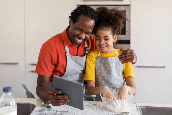 Black Father Baking With Preteen Daughter At Home, Checking Cookies Recipe Online On Digital Tablet, African American Dad And Female Child Using Modern Gadget In Kitchen While Cooking Homemade Pastry