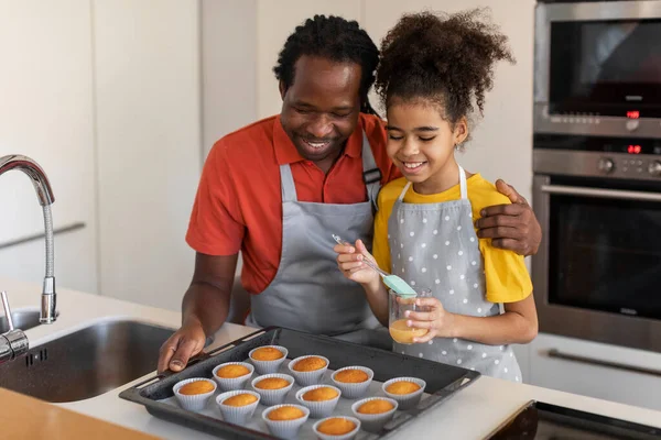 Black Dad And Female Child Baking Muffins Together At Home, Cute Preteen Daughter Brushing Top Of Pastry With Egg Yolks Before Putting It To Oven, Enjoying Making Pastry At Home, Closeup