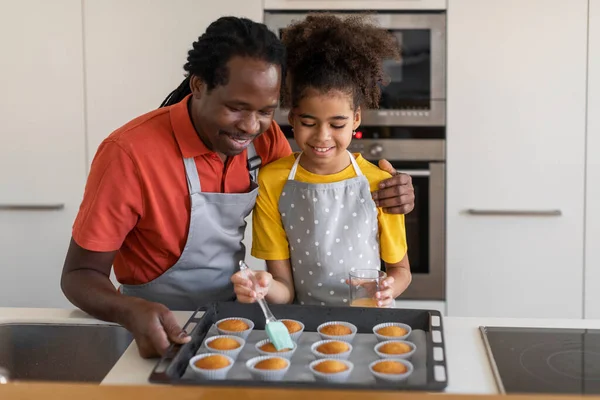 キッチンでお父さんと一緒に焼きながらマフィンに卵の黄身をブラッシングかわいい黒の女の子 家庭で一緒にペストリーを調理しながら幸せなアフリカ系アメリカ人の父と十代の娘を持っています スペースをコピー — ストック写真