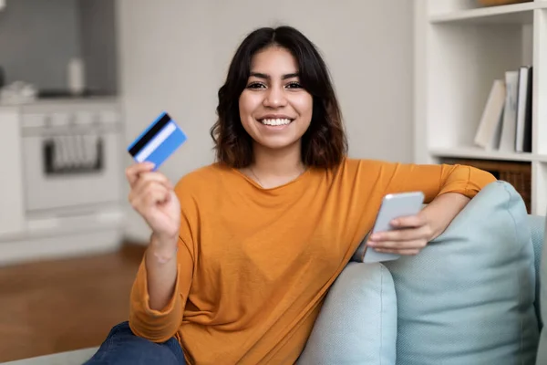 Online Payments. Smiling Arab Female With Smartphone And Credit Card In Hands Sitting On Couch At Home, Happy Young Middle Eastern Woman Making Internet Shopping, Enjoying E-Commerce, Free Space